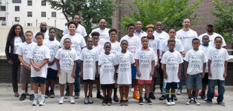 A large group of around 25 Black youth, aged five to 25 stand in a group wearing white T-Shirts featuring the Black Boys Code logo. Behind them is a city building background.