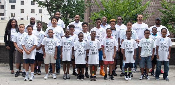 A large group of around 25 Black youth, aged five to 25 stand in a group wearing white T-Shirts featuring the Black Boys Code logo. Behind them is a city building background.