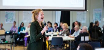 Business owner of Roots and Rivers, Annelies speaking at a workshop holding a microphone with one hand and smiling at the Senior Housing Lab Workshop.