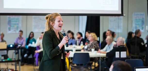 Business owner of Roots and Rivers, Annelies speaking at a workshop holding a microphone with one hand and smiling at the Senior Housing Lab Workshop.