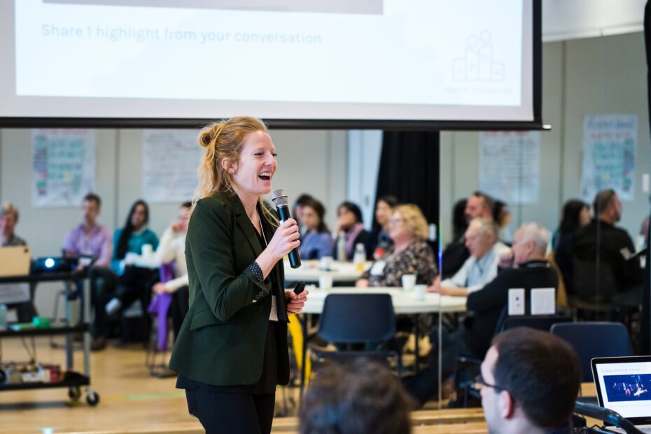 Business owner of Roots and Rivers, Annelies speaking at a workshop holding a microphone with one hand and smiling at the Senior Housing Lab Workshop.