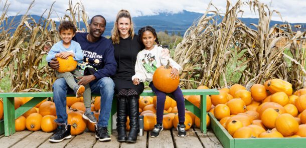 Isaac and Hannah sitting on a green wooden bench next to their children who are each holding a pumpkin with field of corn stalks in the background.