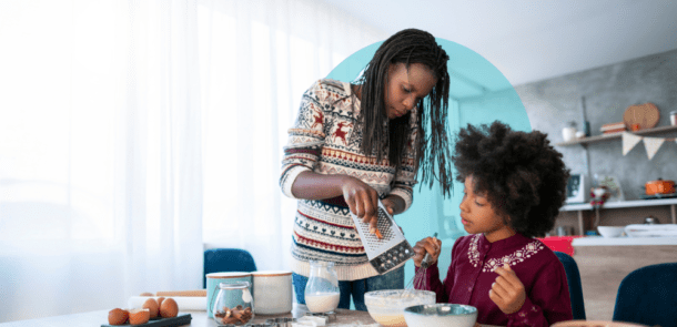 A woman grates an ingredient over a bowl while a child stirs batter in a cozy, well-lit kitchen. The table is filled with ingredients like milk, eggs, and nuts, suggesting they are baking together. The scene conveys warmth, learning, and collaboration.